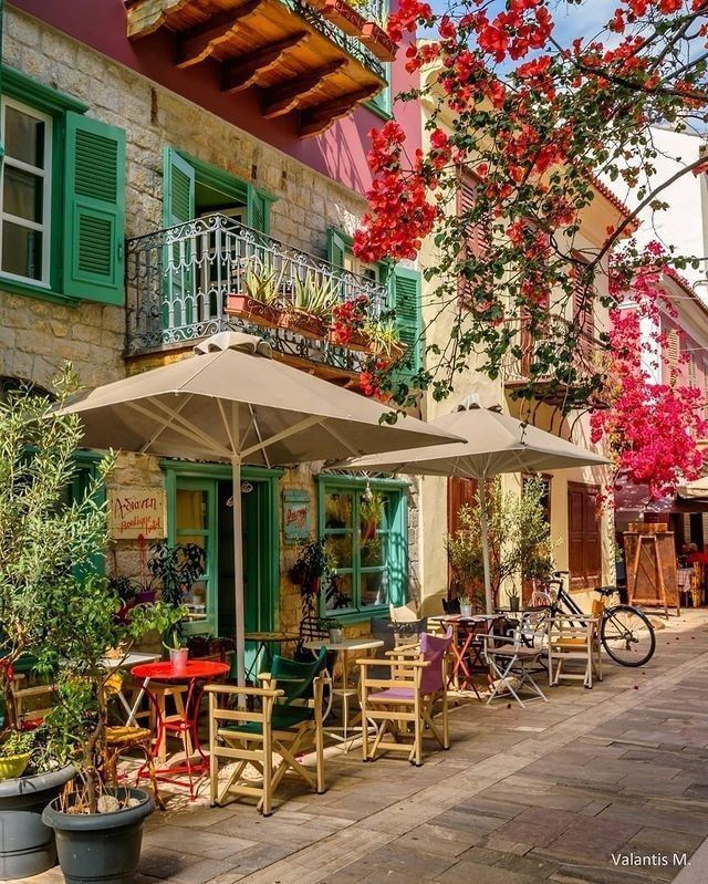 Stone building with green shutters covered in red and pink bougainvillea flowers, cafe tables and bicycles on cobblestone street