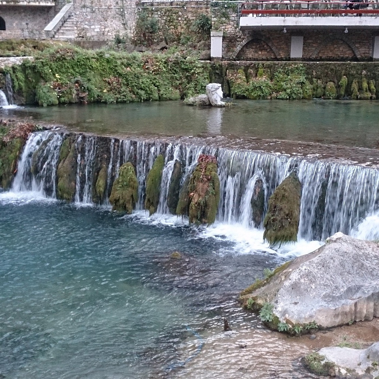 Small river waterfall over a weir with rocks, calm water, and a stone wall behind