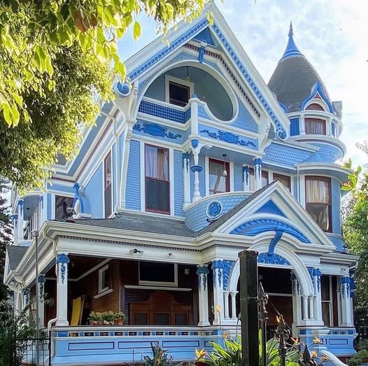 Light blue Victorian house with ornate trim, arched porch, and turret under leafy trees