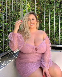 Smiling blonde woman in a sheer purple dress sitting outdoors with greenery behind her