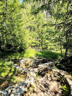 Stone-lined spring along a forested trail with lush green trees and dappled sunlight