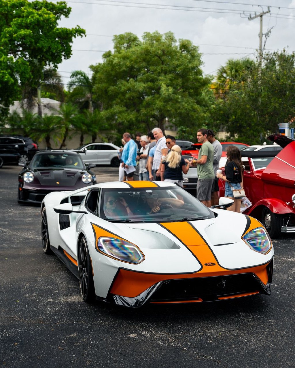 White and orange Ford GT supercar parked at an outdoor car show with people and other cars in the background.