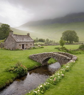 Stone cottage and arched stone bridge over a stream in a misty green valley