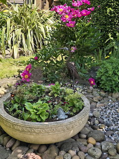 Garden planter with pink flowers, green plants, pebbles, and a small frog ornament surrounded by landscaped garden beds