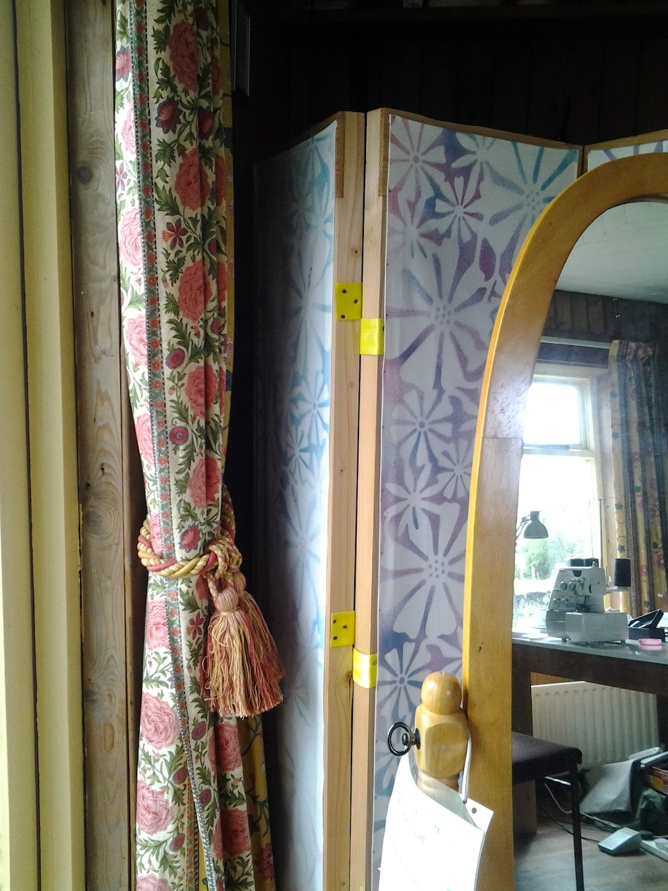 Dressmaker's room with floral curtain, patterned folding screen, and arched mirror reflecting a sewing area