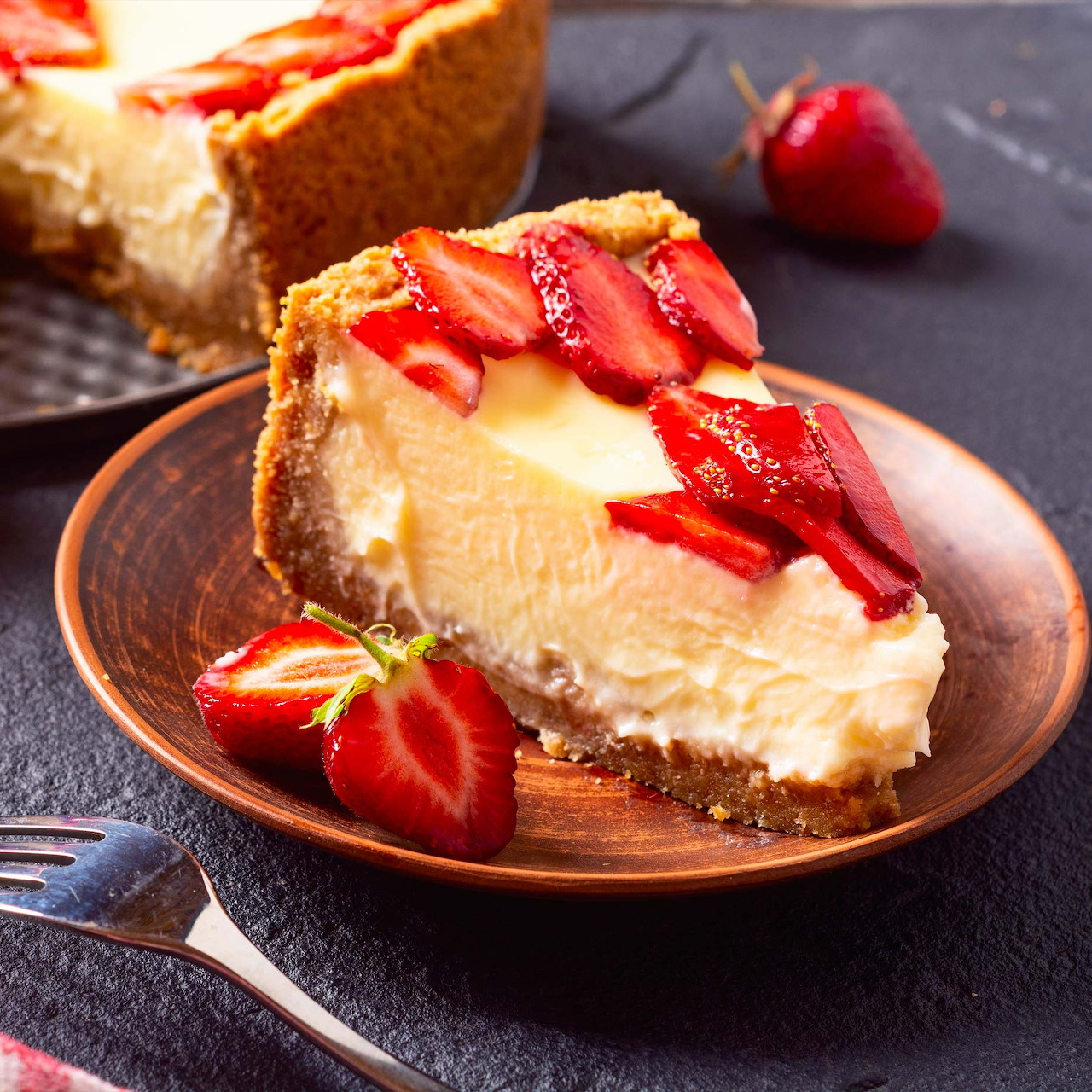 Slice of cheesecake with fresh strawberries on top, served on a brown wooden plate