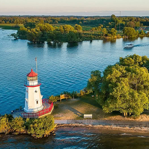 Red and white lighthouse on tree-lined shore with boats on blue water at sunset