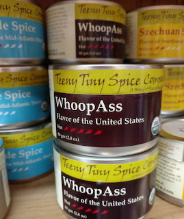 Several small spice cans labeled Whoop Ass from Teny Tiny Spice Company on wooden shelves