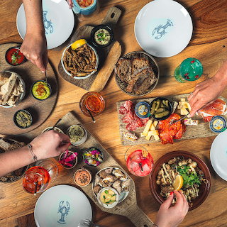 Overhead view of a wooden table with seafood dishes, sauces, plates, and hands serving food