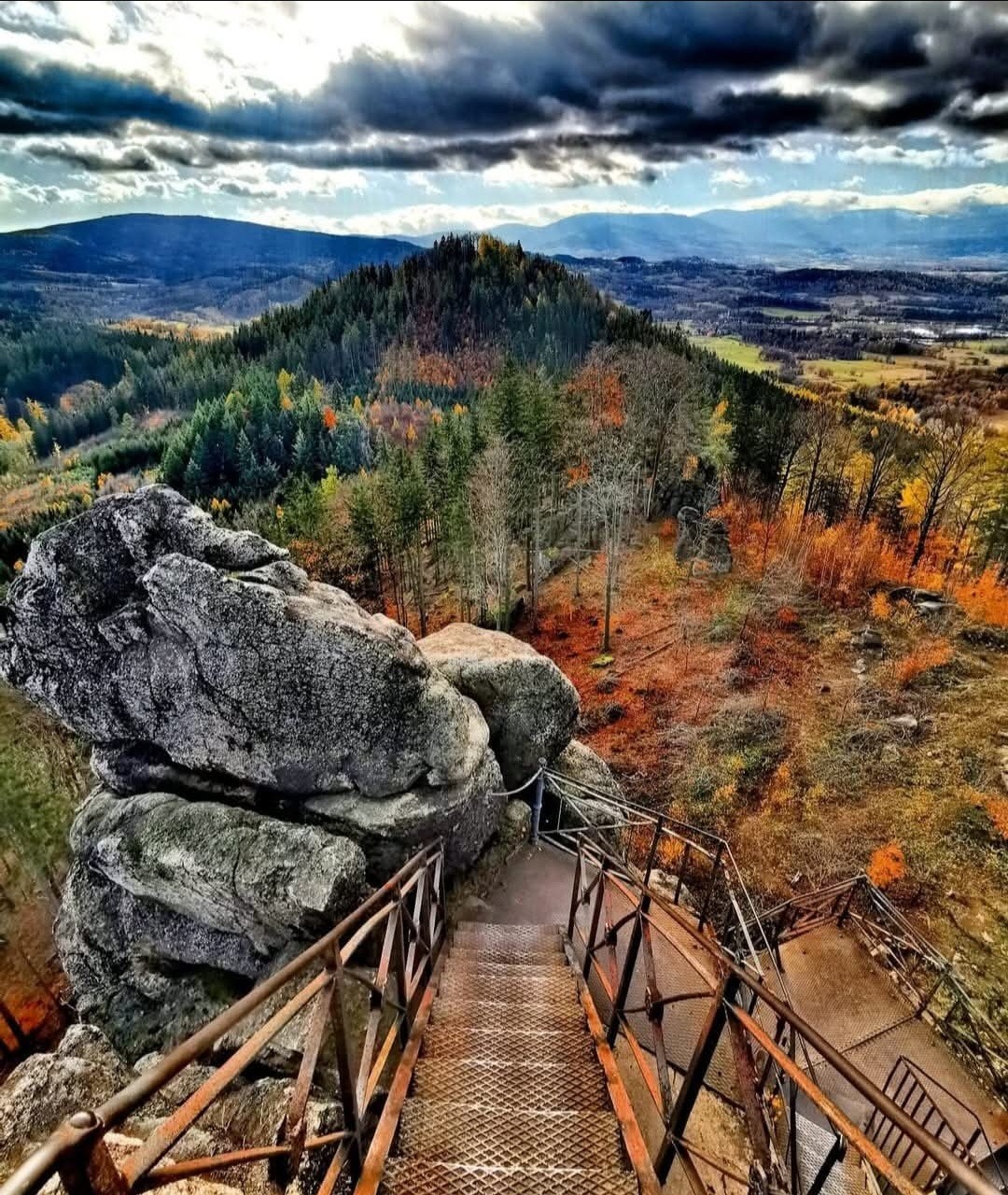 Metal stairs descending from rocky summit with colorful autumn forest valley and dramatic clouds below