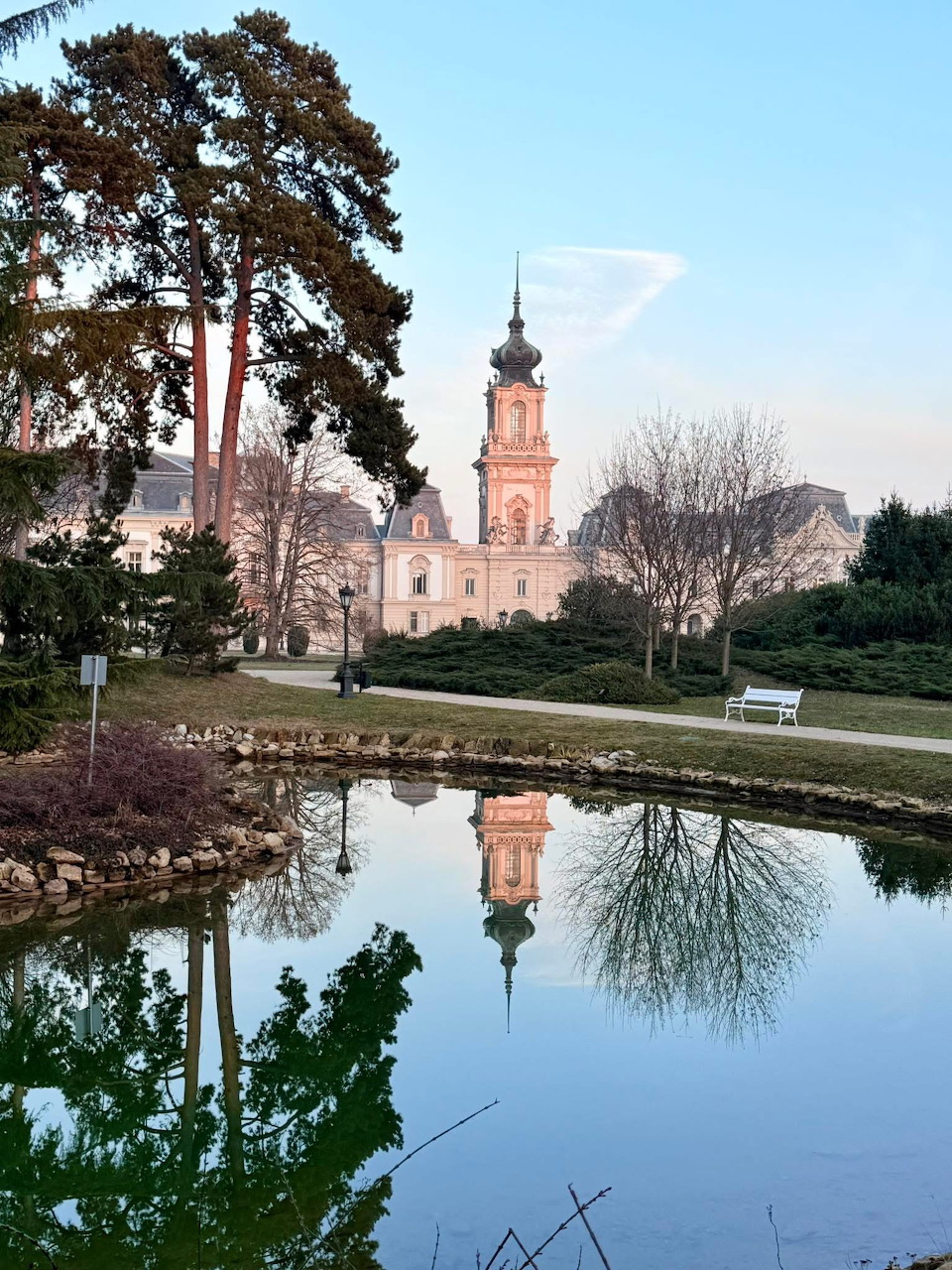 Orange baroque tower reflected in pond at sunset in palace park with trees and lawn