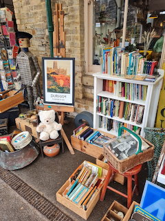 Outdoor vintage shop display with books, framed art, wicker baskets, and collectibles arranged on shelves and crates
