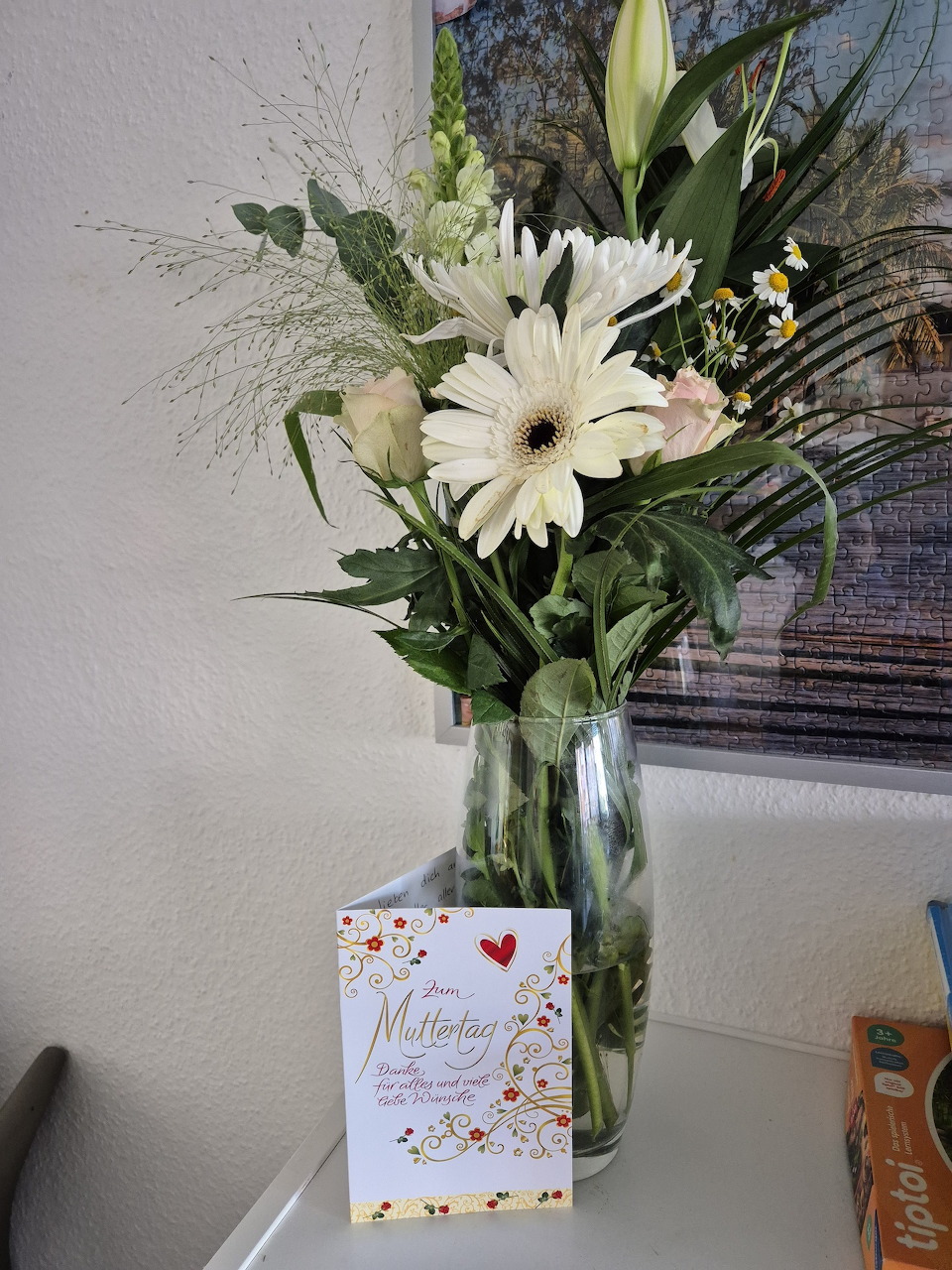 Glass vase with white gerbera daisies and lilies beside a Mother's Day card on a white table