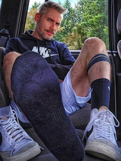 Man in Nike hoodie, dark socks, shorts, and white sneakers sitting inside a car with legs extended