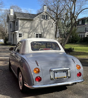 Light blue Nissan Figaro retro car parked on a driveway with a white house and trees behind it