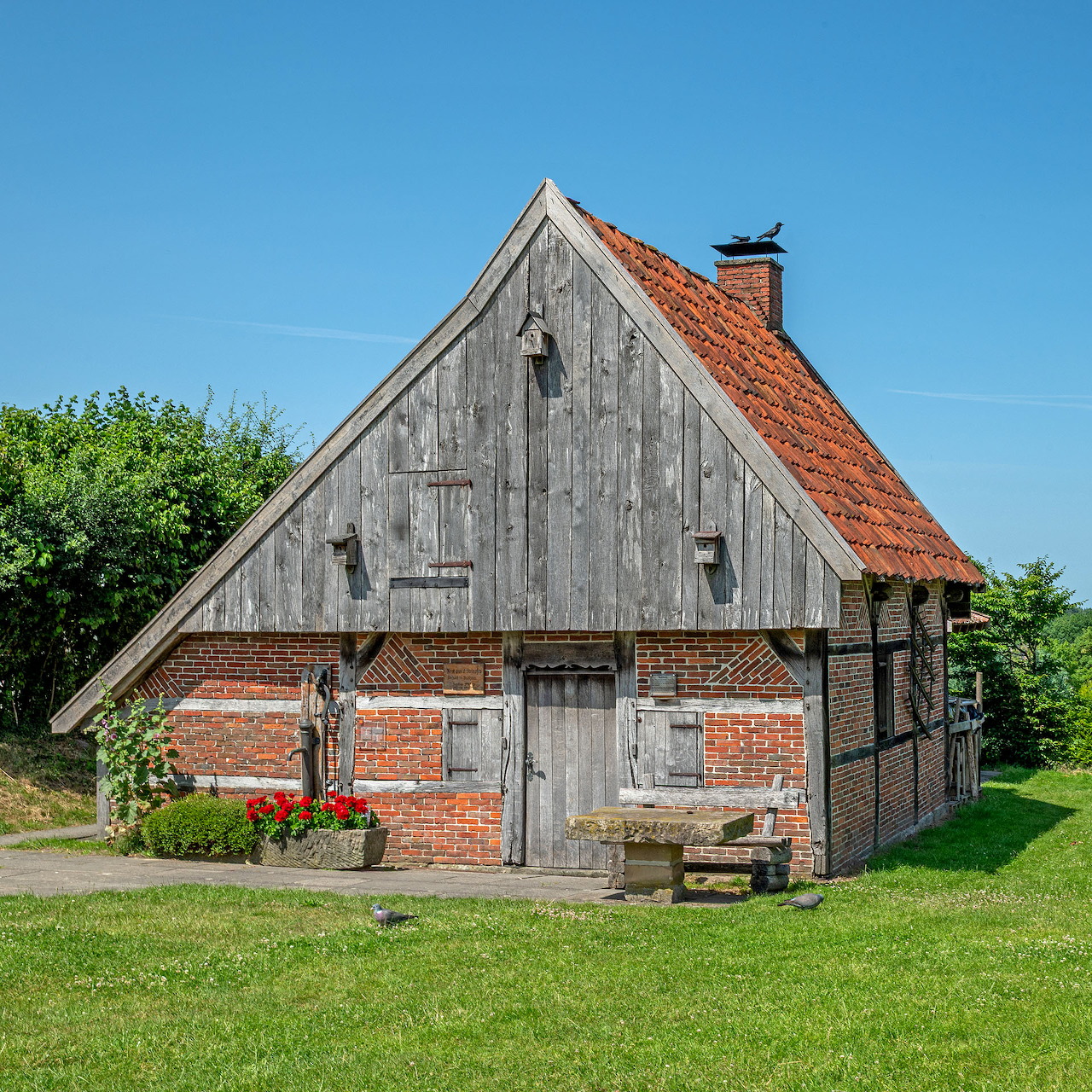 Rustic wooden barn with red brick foundation and orange tile roof on green grass under blue sky
