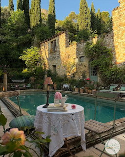 Stone Mediterranean villa with ivy overlooking a turquoise pool, white table in foreground, cypress trees