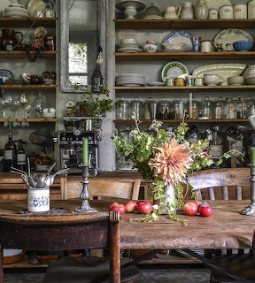 Rustic kitchen with open wooden shelves, vintage dishes, farmhouse table, and fresh flower arrangement