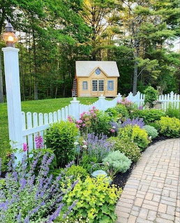 Miniature yellow cottage behind white picket fence with colorful flower garden and brick path
