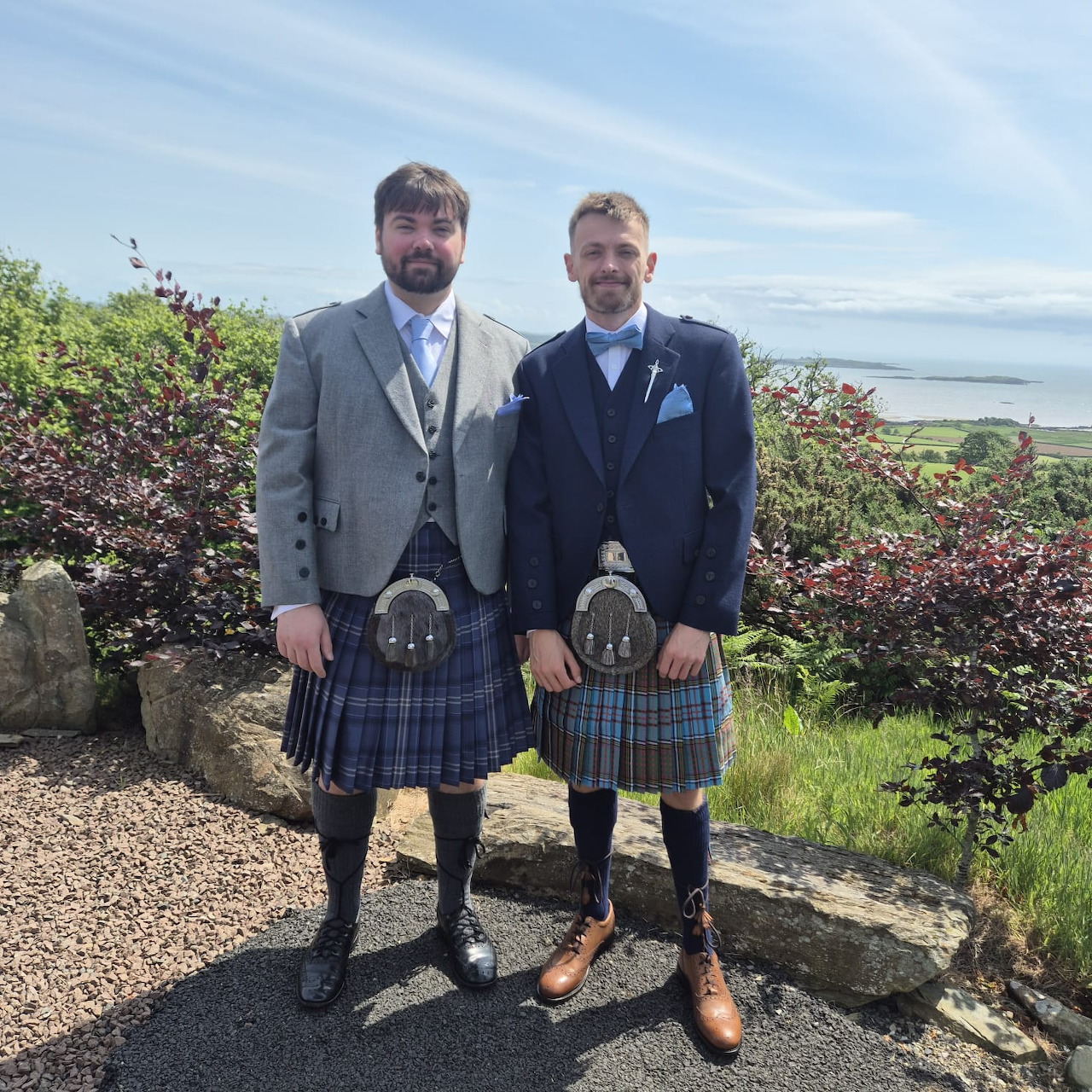 Two men in formal jackets and tartan kilts standing outdoors with a coastal view behind them
