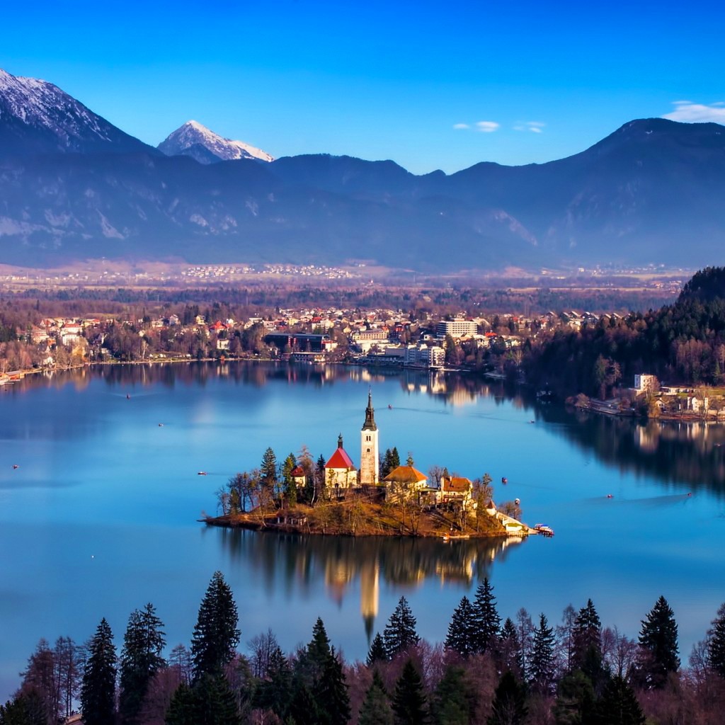 Small island with white church and tower in blue lake, mountains and town in background, twilight sky