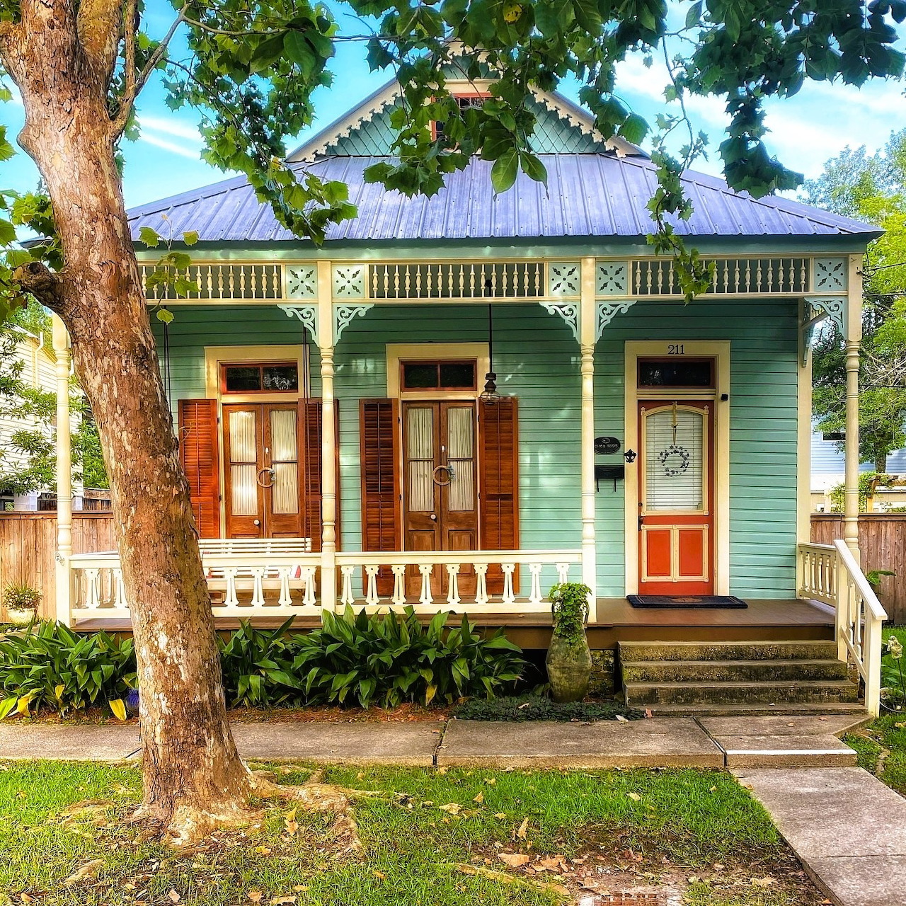 Green Victorian cottage with tin roof, white porch railing, orange door, and large tree in front yard