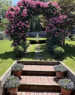Lush garden with pink rose archway over fountain and brick pathway