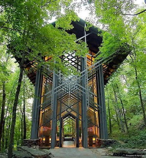 Glass and wood chapel with geometric lattice structure surrounded by lush green forest trees