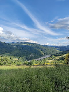 Grassy hillside overlooking a green valley and forested hills under a blue sky with wispy clouds.