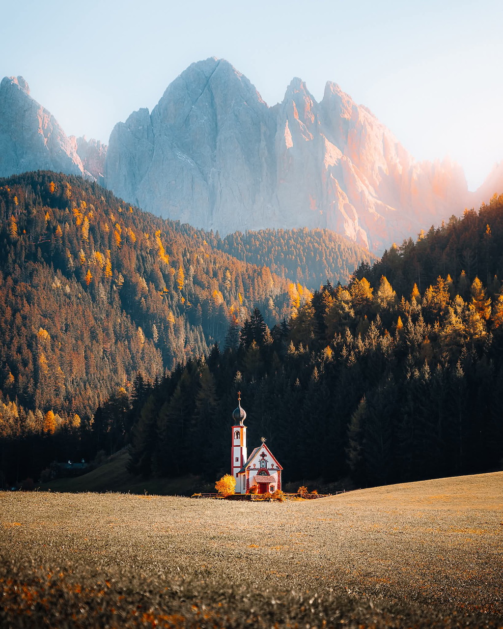 Small white church in a meadow surrounded by autumn trees and jagged Dolomite mountain peaks