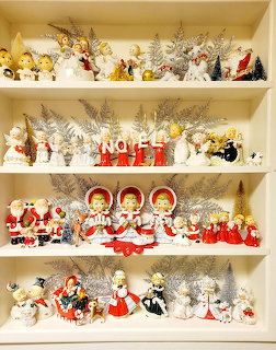 Four white shelves filled with vintage Christmas figurines, angels, Santa Claus figures, and holiday decorations