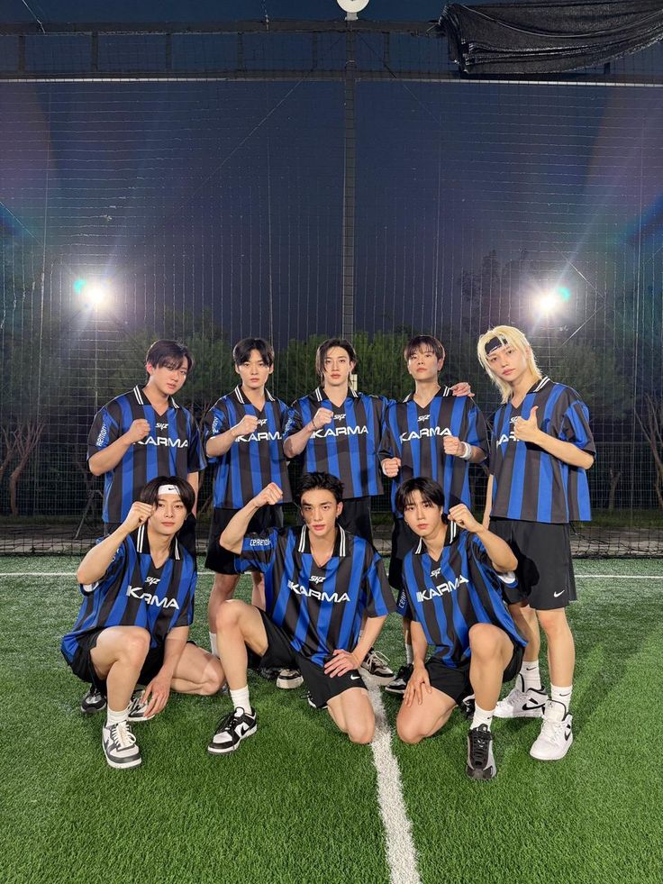 Nine young men in matching blue and black soccer jerseys posing on an indoor turf field at night.