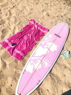 Pink surfboard with white flowers lying on sand beside magenta towel and flip-flops