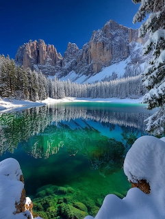 Crystal-clear turquoise alpine lake reflecting snowy mountains and frost-covered pine trees in winter