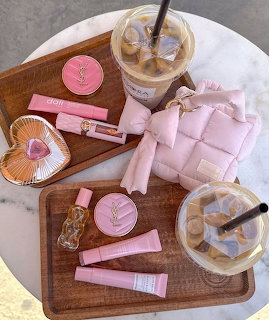 Pink makeup items, quilted pink bag, and iced coffees on wooden trays on a marble table