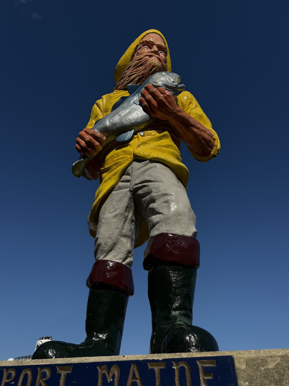 Painted fisherman statue in yellow rain gear holding a fish, Port Maine sign on base, blue sky background