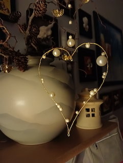 Wire heart shape adorned with white pearls displayed on a wooden shelf next to a round vase and small ceramic house