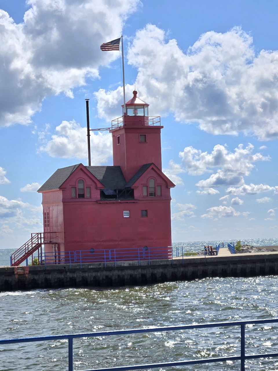 Red lighthouse on a concrete pier with American flag on top, lake water and cloudy sky in background
