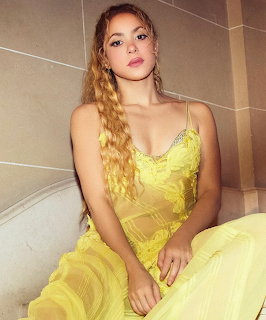 Woman with long wavy blonde hair wearing a yellow lace dress sitting against a tiled wall