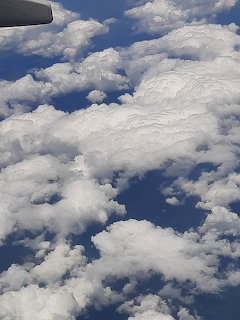Aerial view of white clouds over blue sky with a small airplane wing visible at top left.