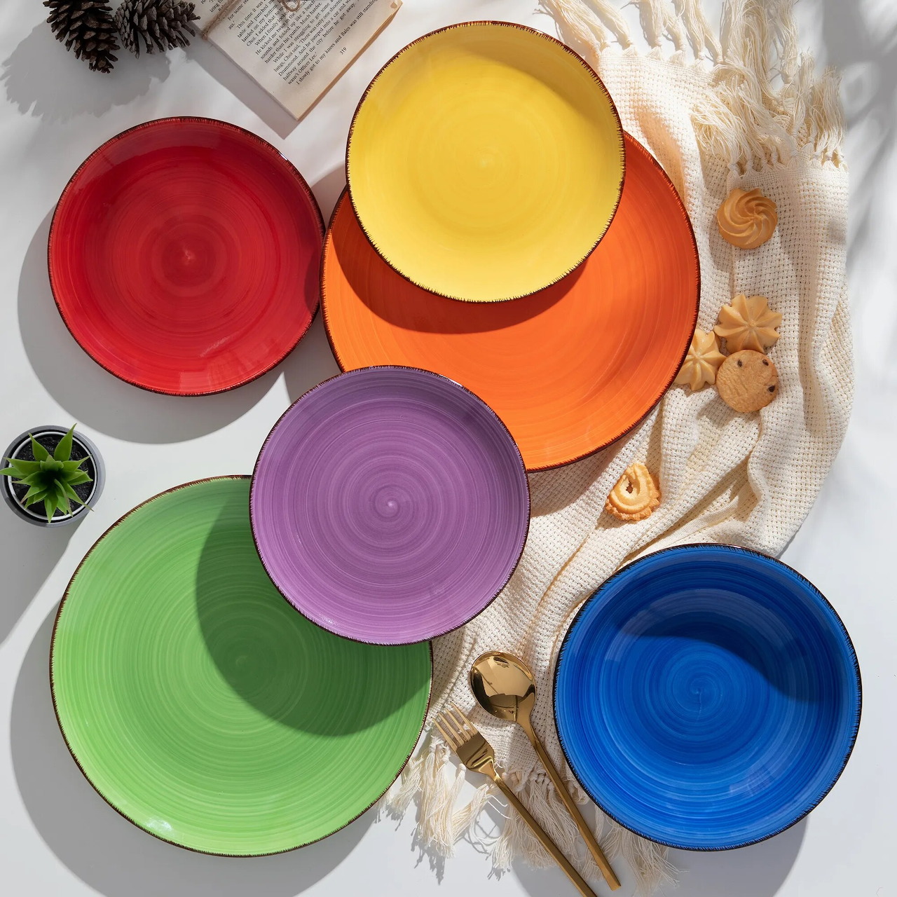 Overhead flatlay of red, orange, yellow, purple, green, and blue ceramic plates on white fabric with dried flowers