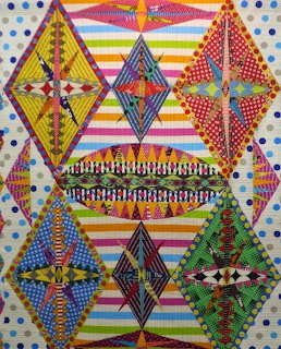 Colorful patchwork quilt with six geometric shield blocks on rainbow striped background and polka dot border