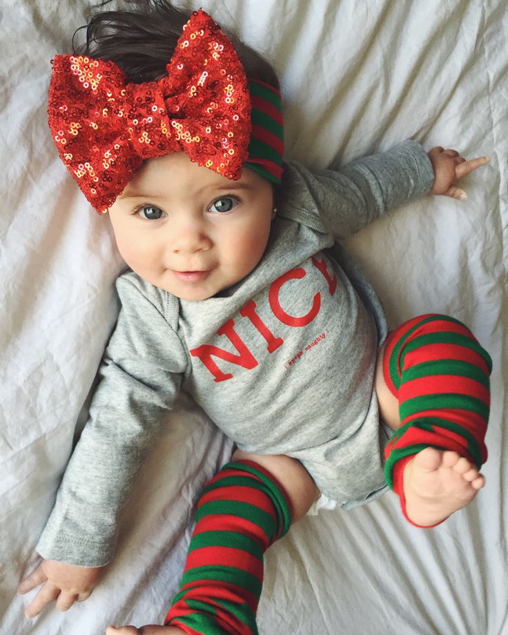 Baby with red sequined bow headband lying on white bedding wearing gray shirt and striped leg warmers