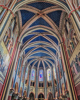 Gothic church interior with ornate blue vaulted ceiling, ribbed arches, stained glass windows, and decorated columns