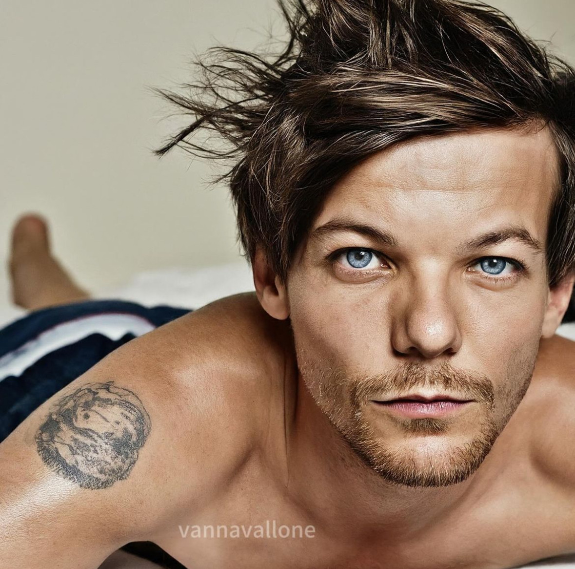 Close-up of a shirtless man with blue eyes, messy hair, and a shoulder tattoo lying on a bed