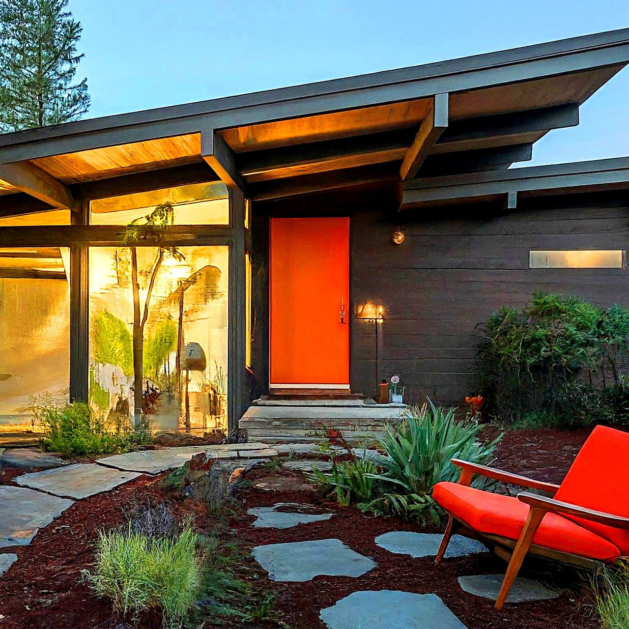Modern home entrance with orange door, exposed wooden beams, stepping stones, and outdoor chair at twilight
