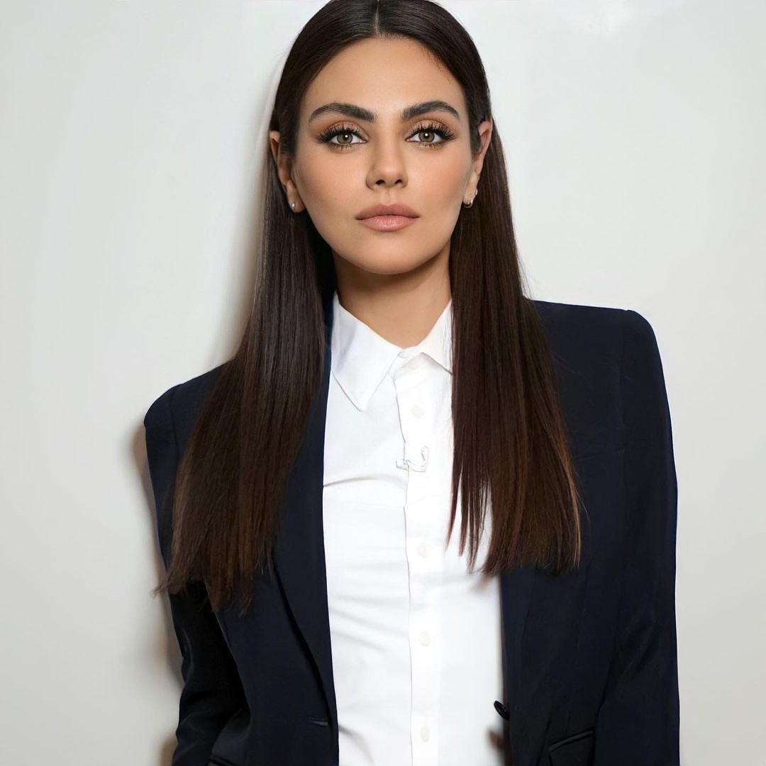 Woman with long straight brown hair in navy blazer and white shirt posing against light background
