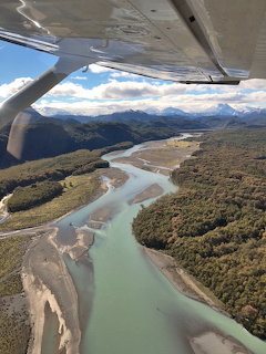 Aerial view from small aircraft over turquoise river flowing through green forested valley with mountains