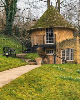 Round stone cottage with conical moss roof and wooden door on green lawn with stone path