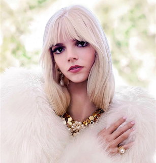 Portrait of Anya Taylor-Joy in a white fur-like wrap with blonde hair and gold jewelry
